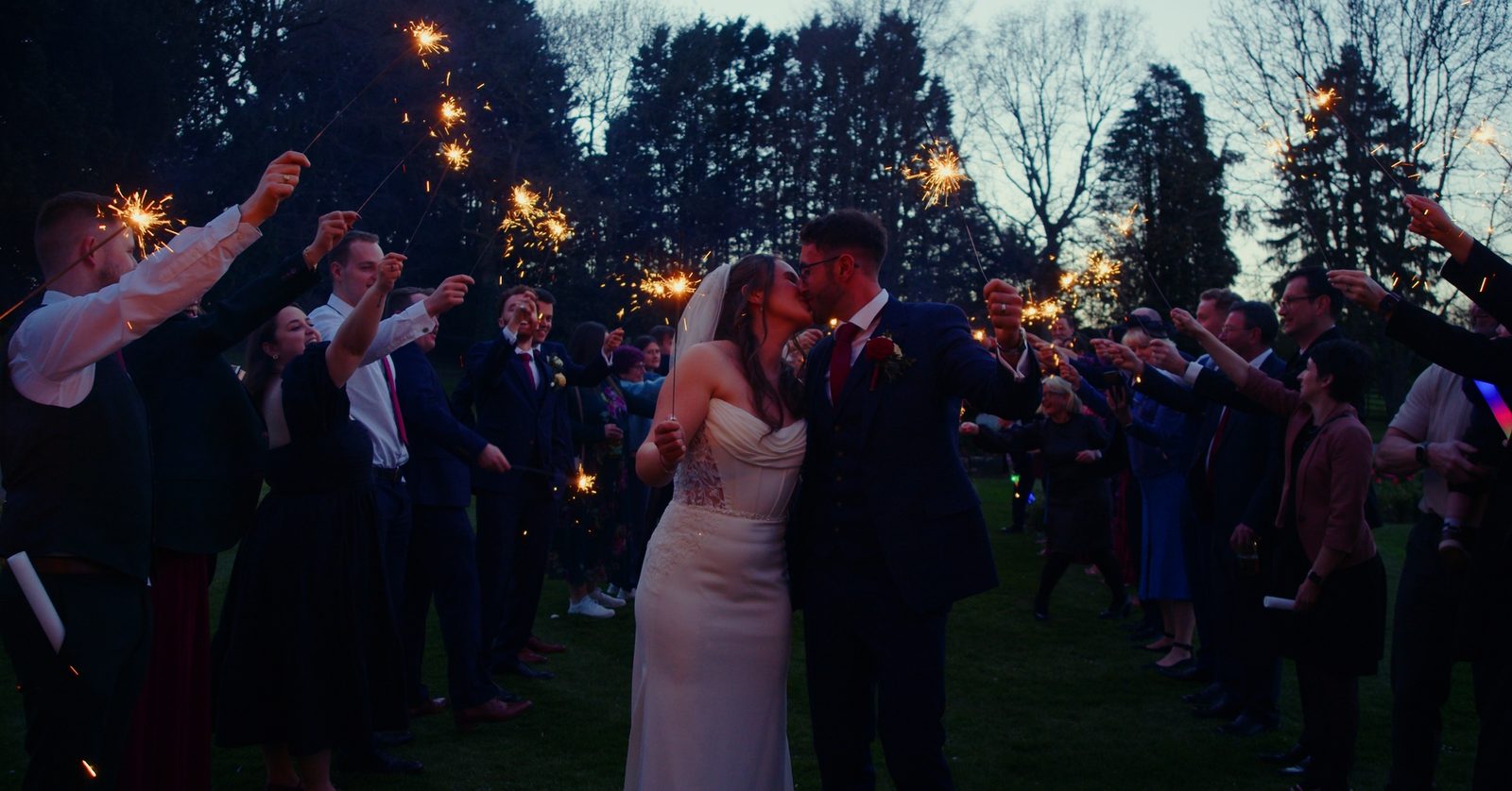 Sarah and Ben kissing beneath an arch of sparklers held by their guests at dusk